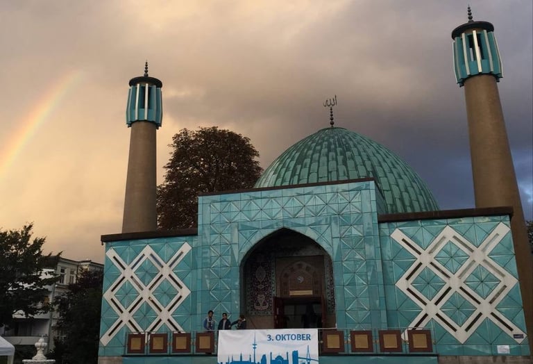 Ornate mosque with turquoise dome, two minarets, geometric tile patterns, and rainbow in sky at dusk